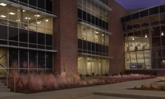 Modern office building with glass curtain walls and brick exterior at dusk, illuminated interior visible through windows.