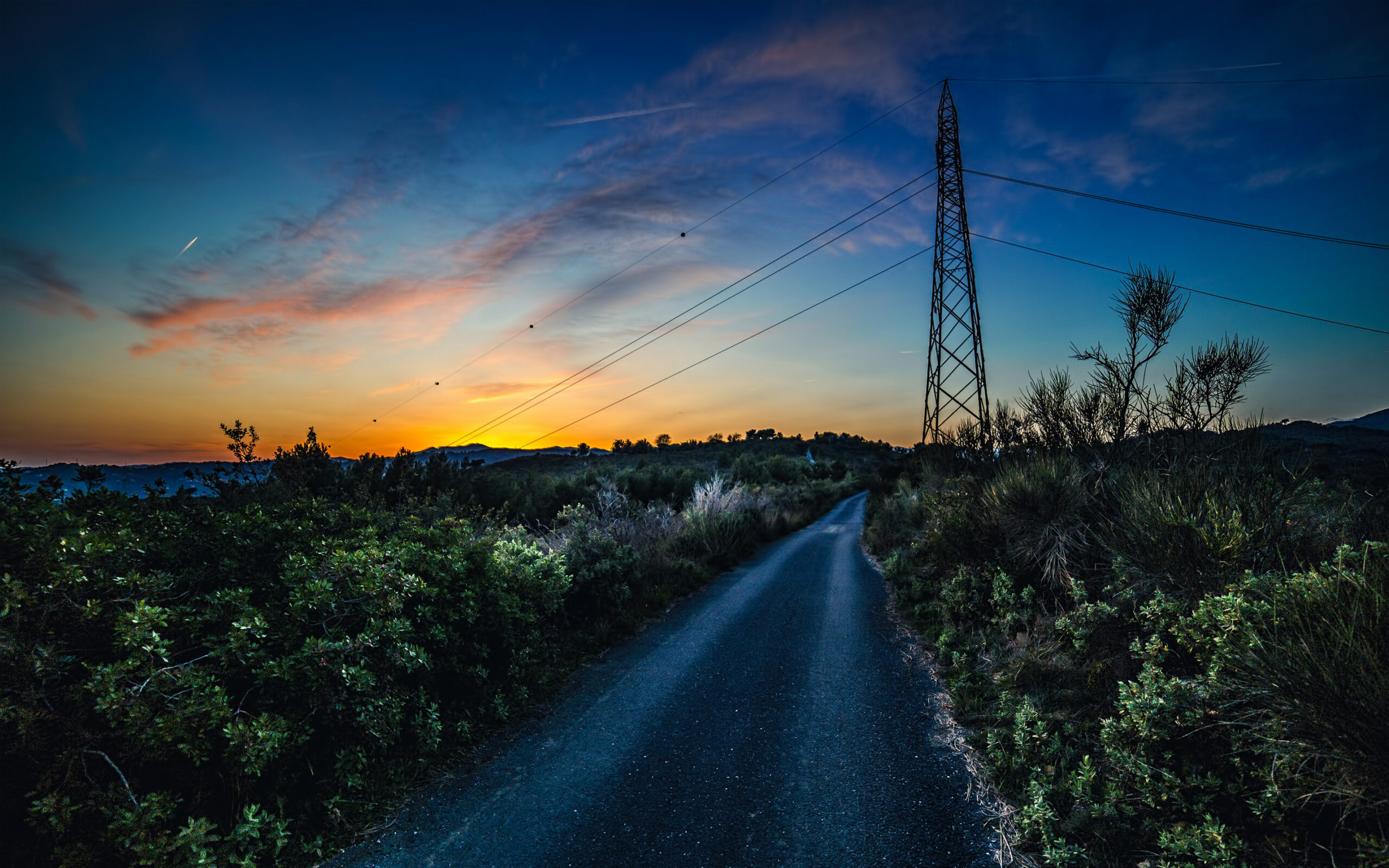 Narrow rural road at sunset with power lines stretching across the sky and a tall transmission tower beside shrubs and hills.
