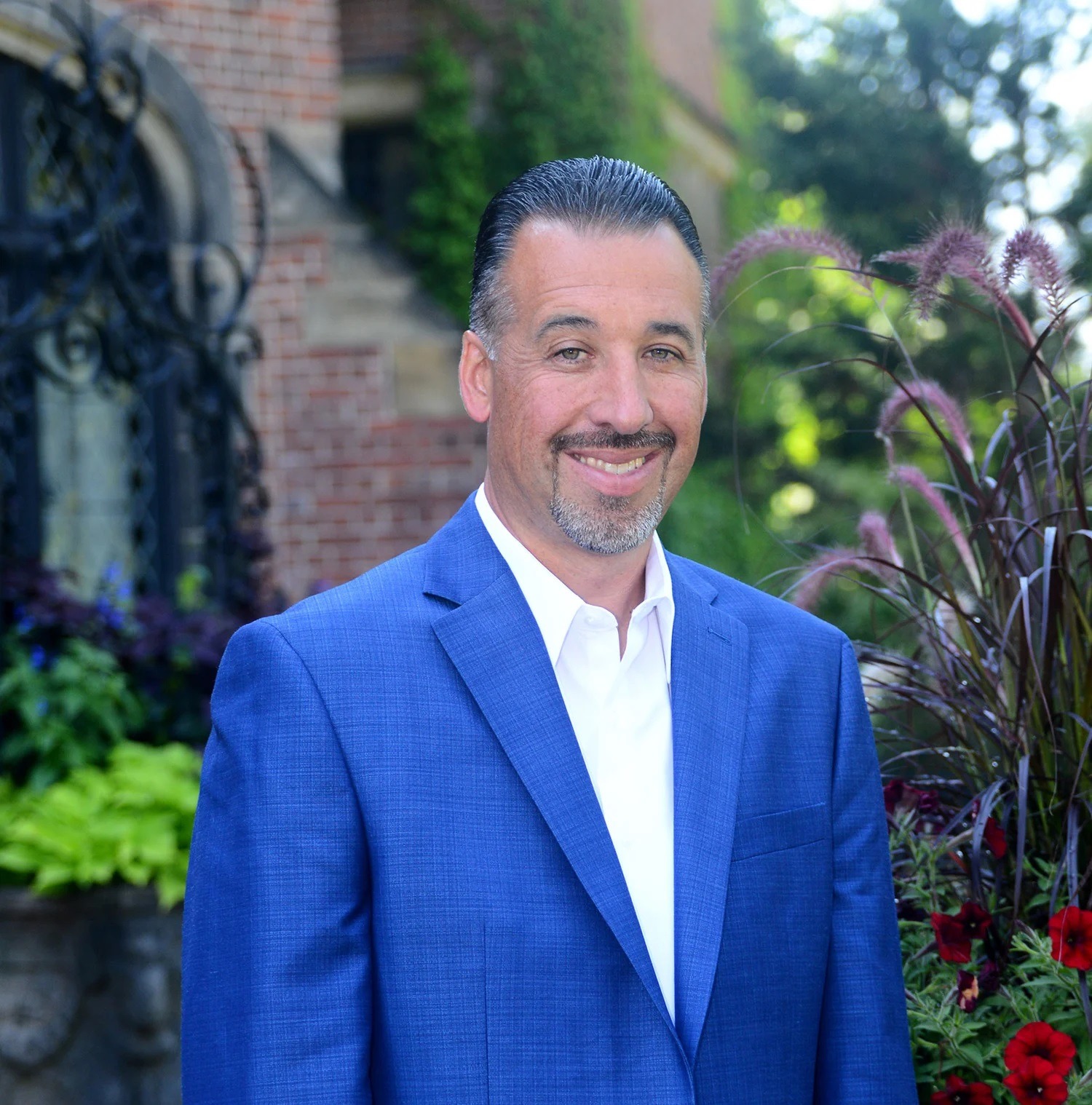 Smiling man wearing a blue blazer and white shirt standing outdoors in a landscaped garden setting.