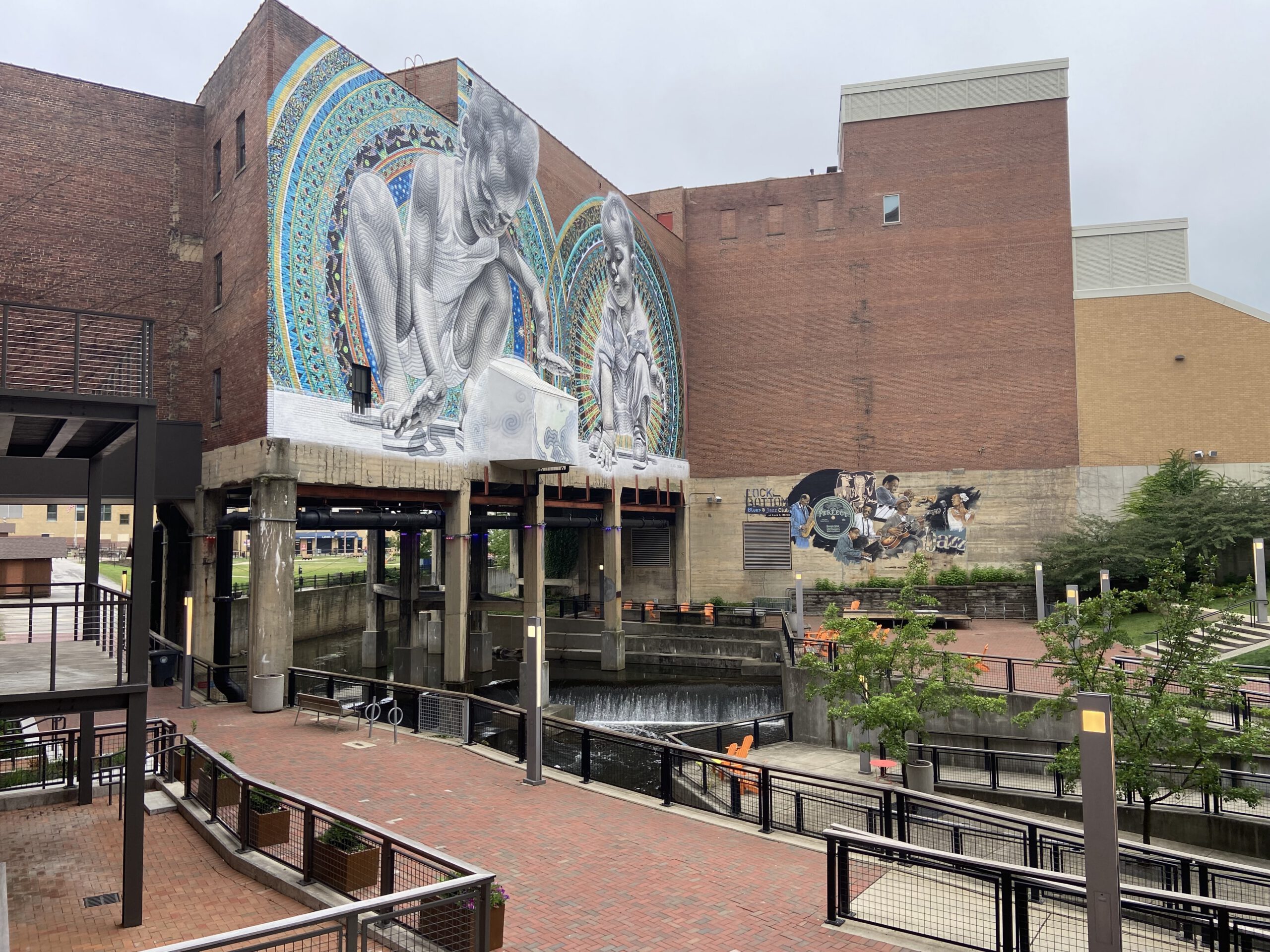 Large mural depicting two kneeling figures on the side of a brick building overlooking a landscaped canal walkway with railings, seating, and water features.