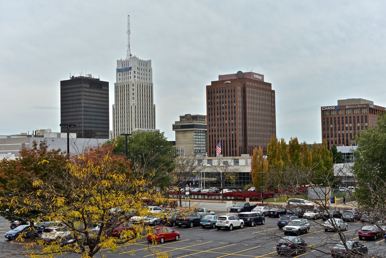 Downtown city skyline with mid-rise office buildings, autumn trees, and a parking lot in the foreground.