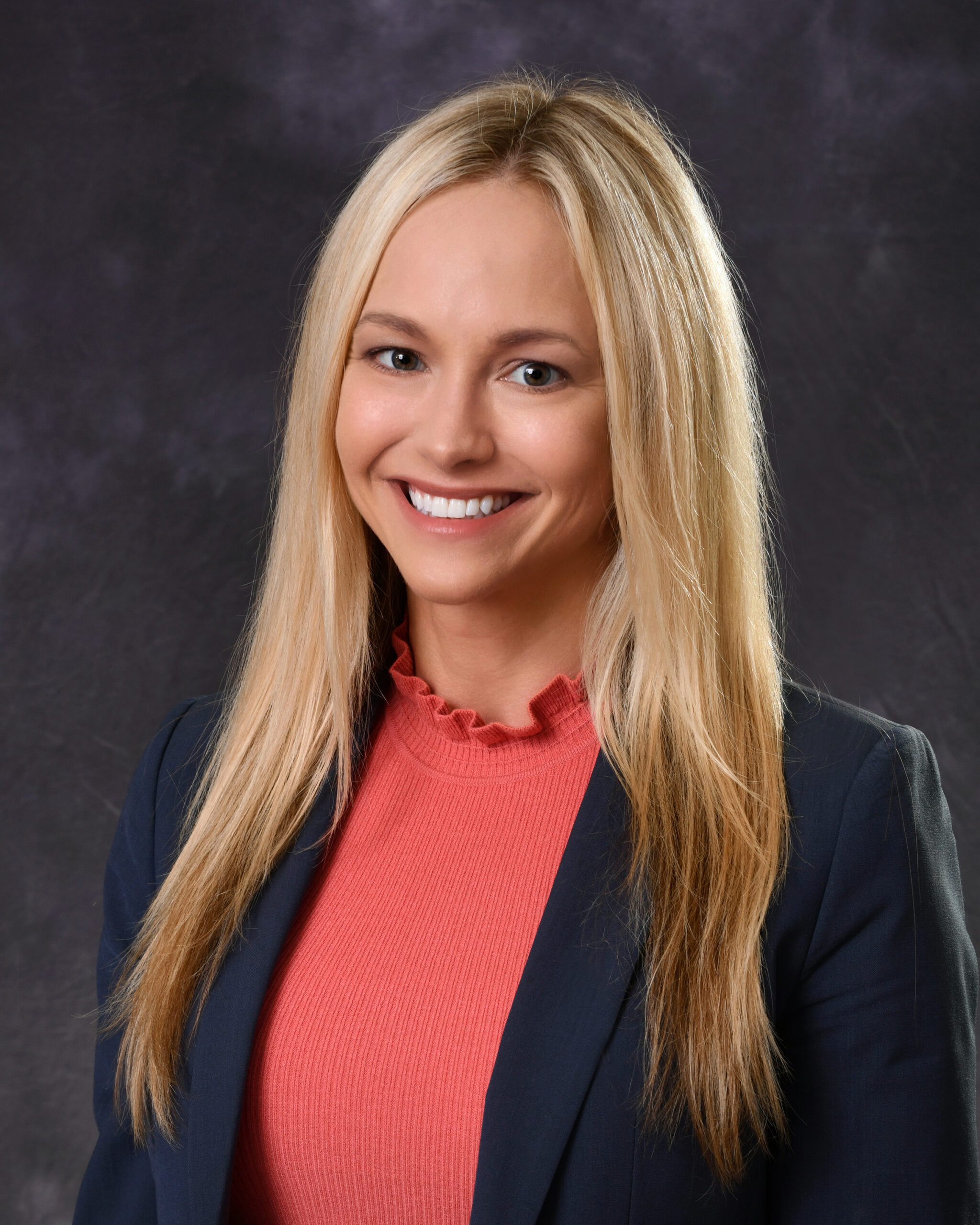 Professional headshot of a smiling woman with long blonde hair wearing a coral top and dark blazer against a studio backdrop.