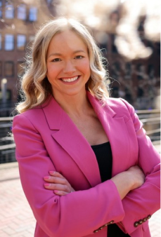 A woman in a pink blazer stands outdoors with arms crossed, smiling confidently in a city setting.