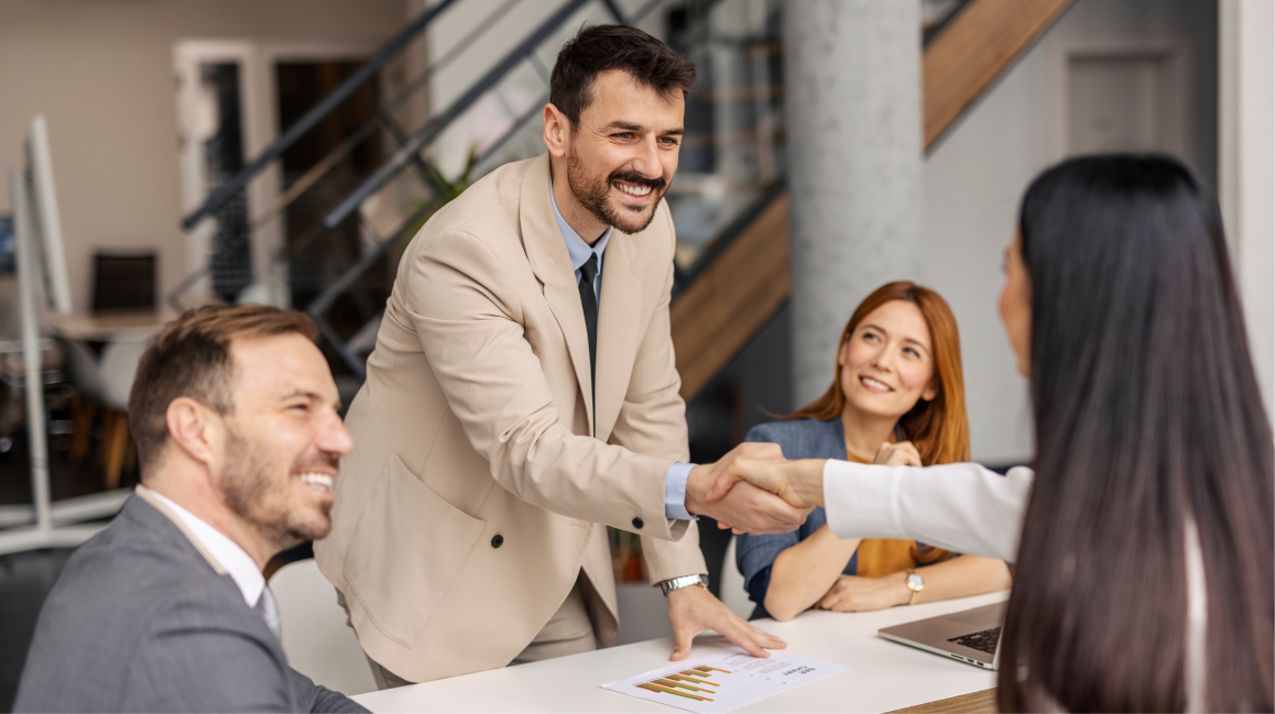 Business professional leaning forward to shake hands during a meeting while colleagues look on.