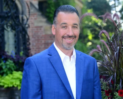 Smiling man wearing a blue blazer and white shirt standing outdoors in a landscaped garden setting.
